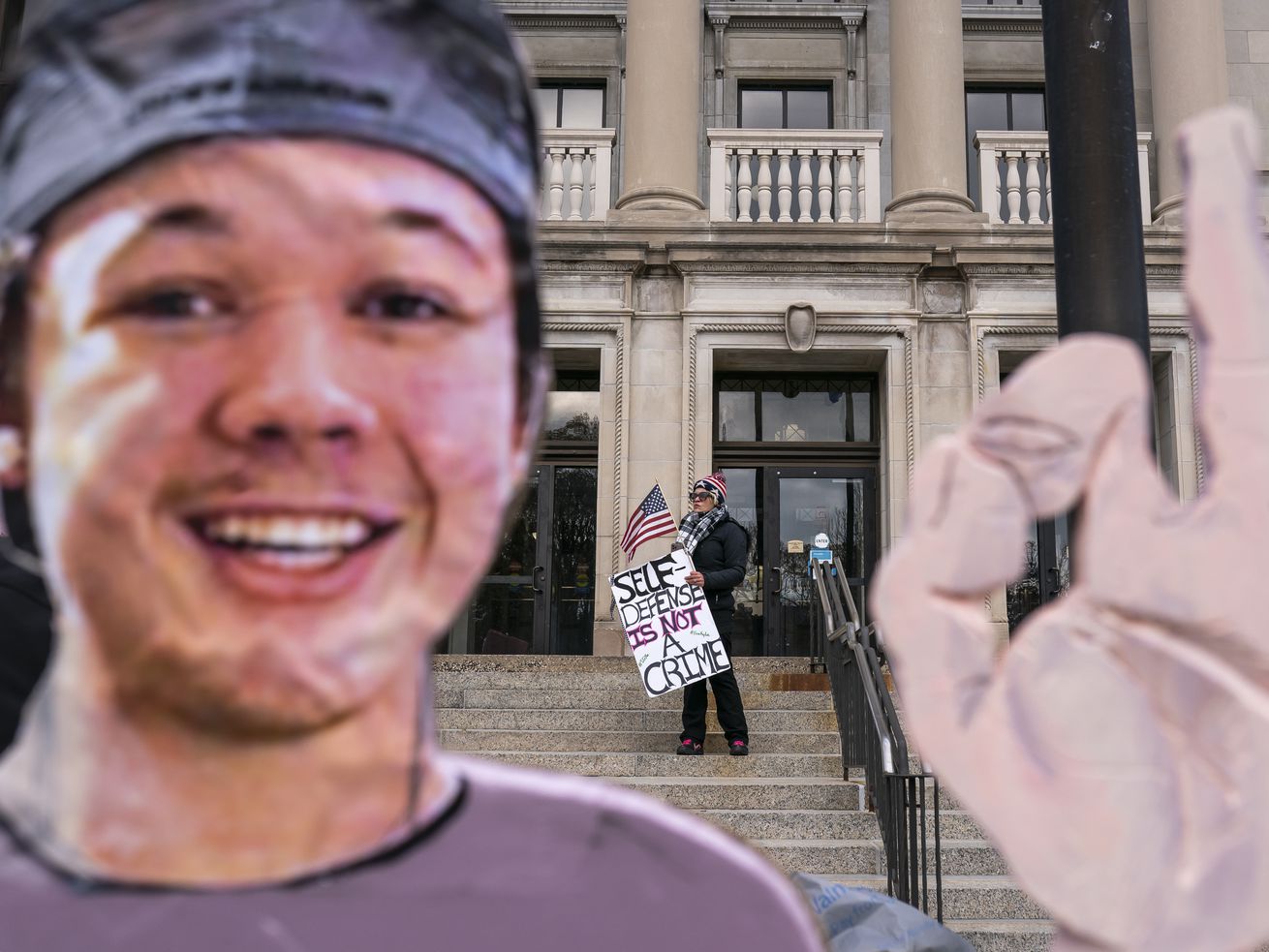 A cut-out of Kyle Rittenhouse’s face and hand in the foreground, the courthouse and a protester in the background.