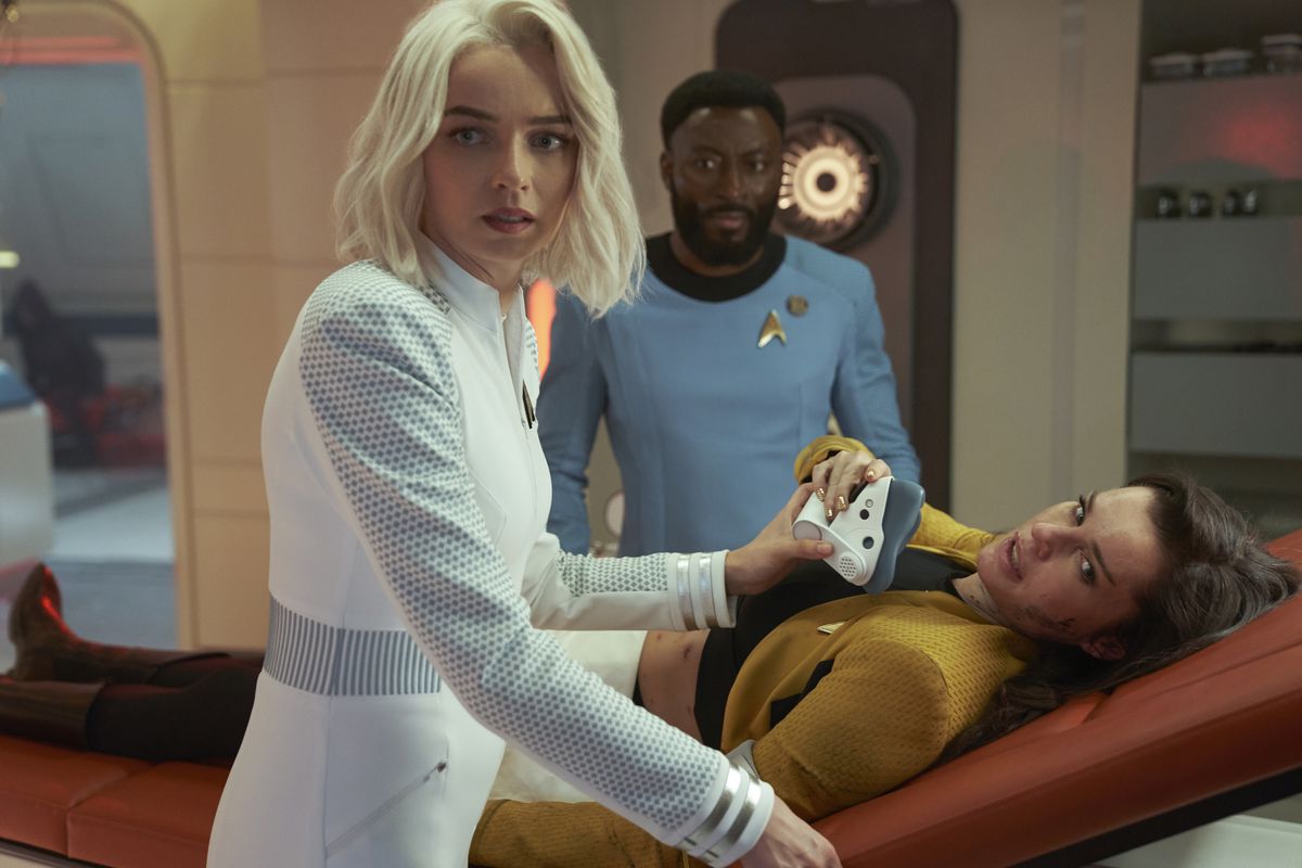 A dark-haired white woman lying on a stretcher is being checked for infection by a blond white woman, and a dark-haired black man looks on anxiously.