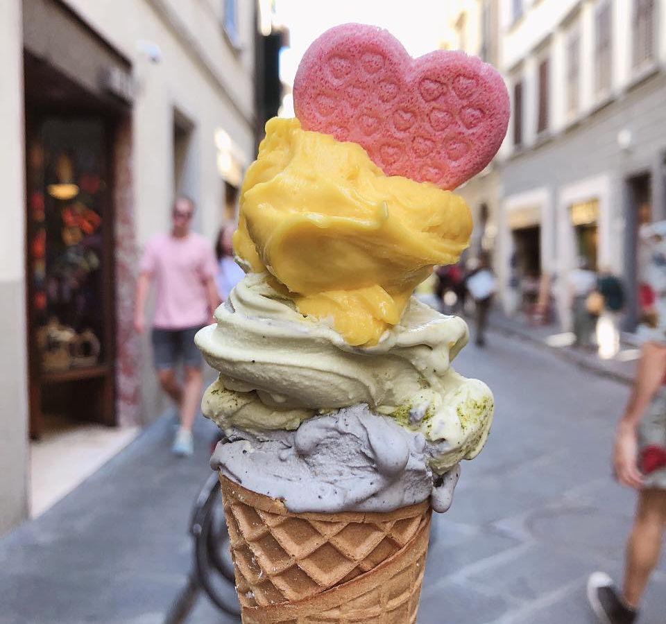 A tilting stack of gelato on a cone topped with an edible heart, in front of a street filled with people