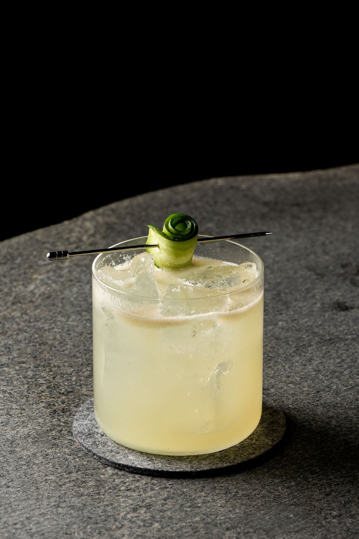 A light yellow cocktail with a swirl of cucumber atop.