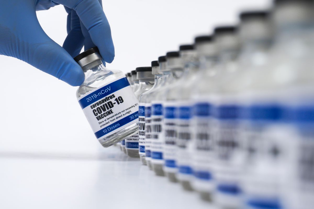 A blue-gloved hand pulls a COVID vaccine-labeled vial from a row of similar vials.
