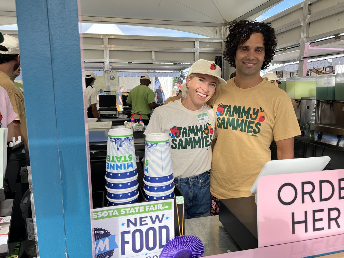 Two people wearing T-shirts smile at the camera in a food stand in front of an “order here” sign. 