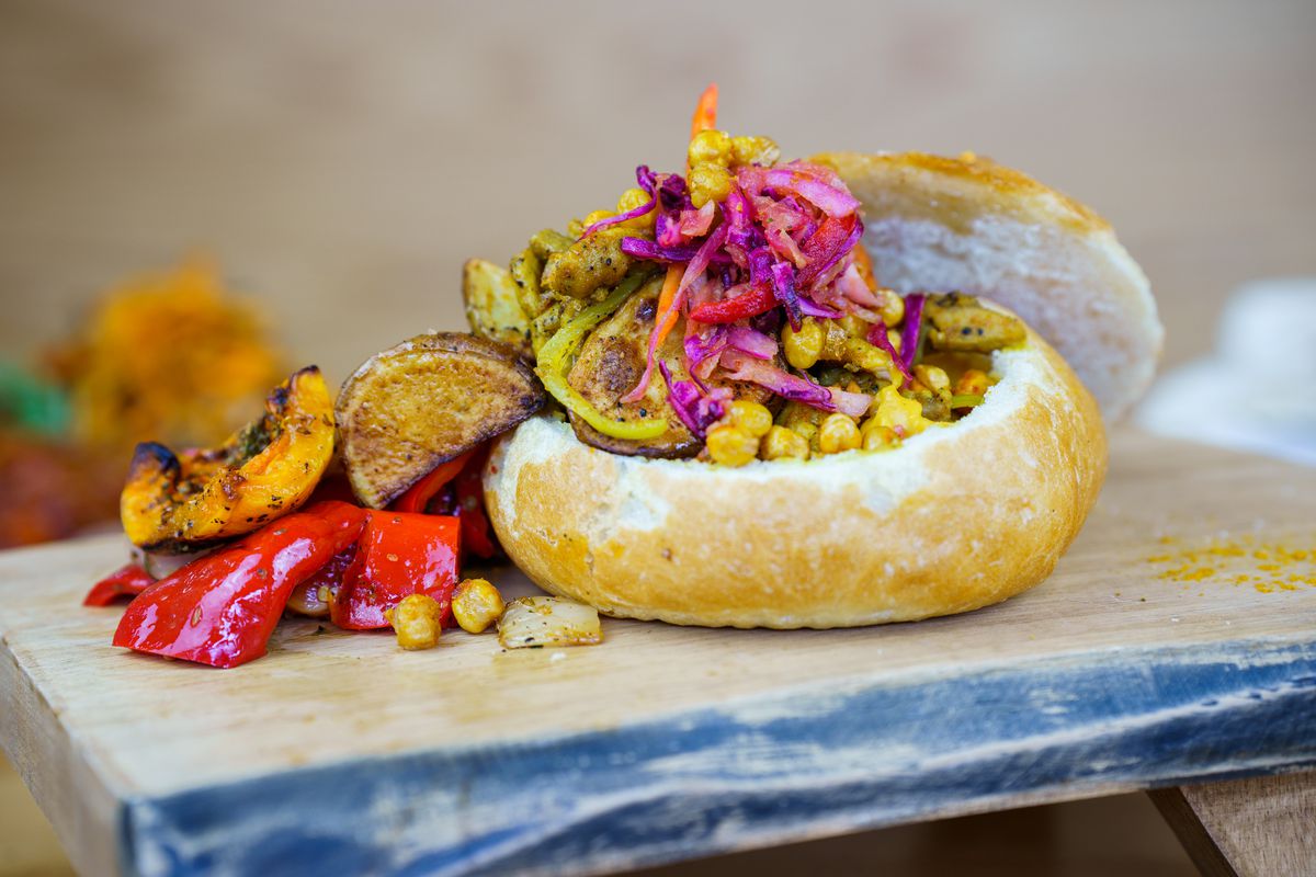 A bread bowl filled with chickpeas, chicken, and vegetables on a wooden platter.