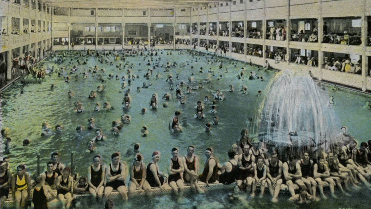 A close-up of a colored vintage postcard showing young men and women crowded inside a massive swimming pool in the Redondo Beach Bath House. There’s a large fountain in the forefront and the pool is rimmed in observation areas.&nbsp;