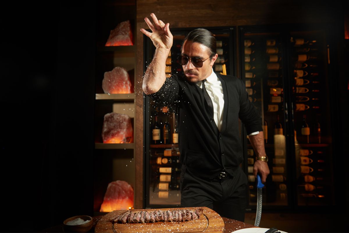 A man in dark sunglasses in a signature salt-sprinkling post stands over a cutting board with sliced steak.