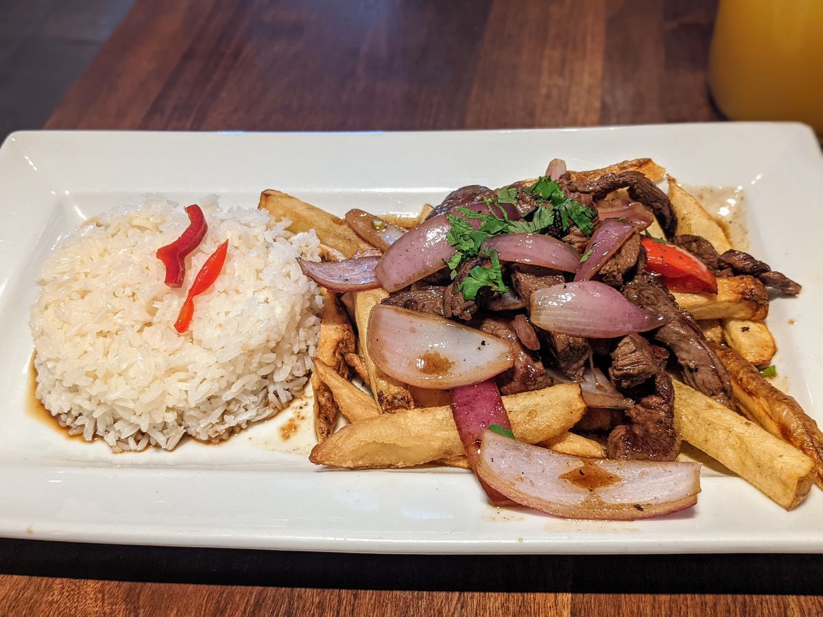 A plate of beef, fries, and rice on a wooden table under harsh light.