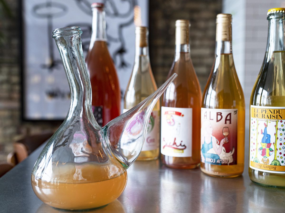 Five bottles of gold, orange, a pink natural wines sit on a table behind a carafe with orange wine in it. 