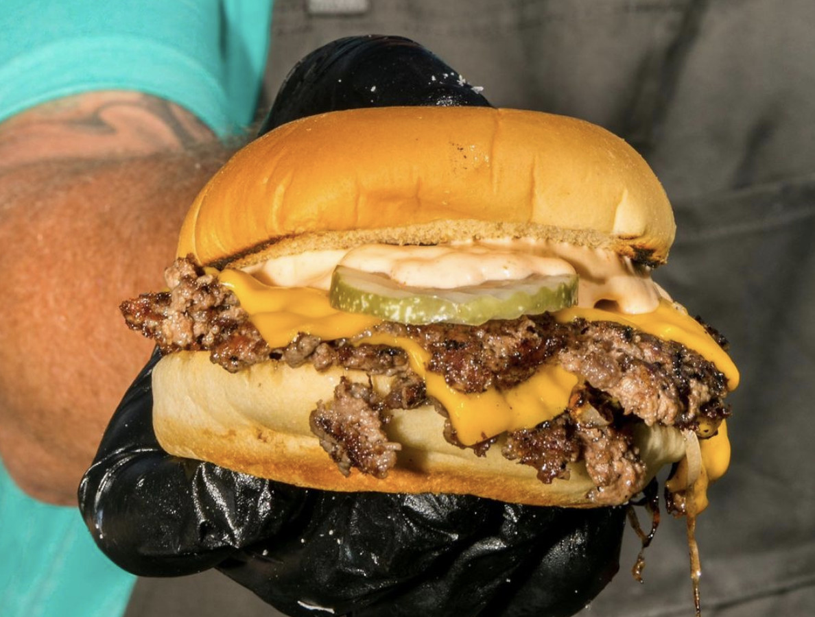 A hand in a black glove holds out a messy cheeseburger.