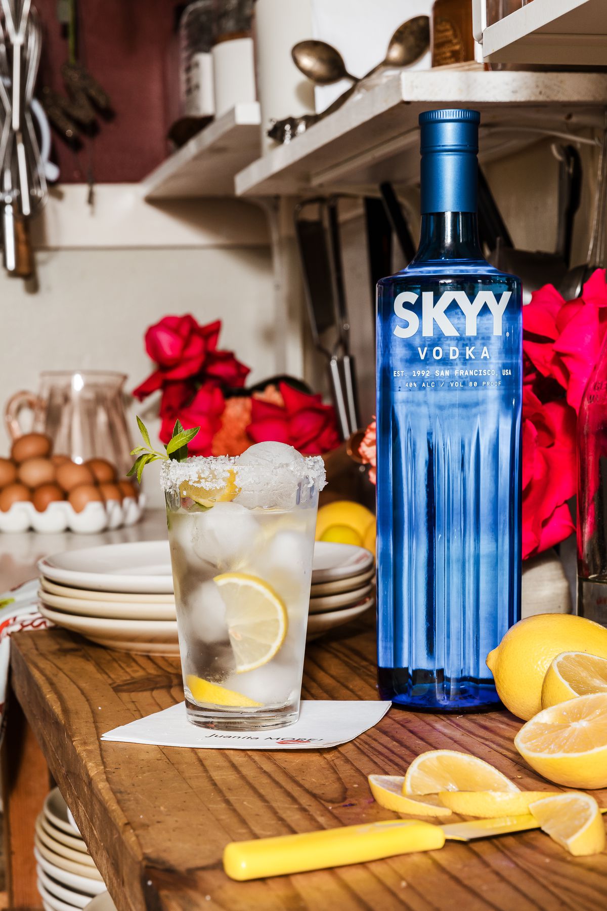 On a countertop is a vodka soda highball, topped filled with sliced oranges, a salted rim, and a sprig of mint as a garnish. Alongside it is a SKYY Vodka bottle along with other sliced lemons. 
