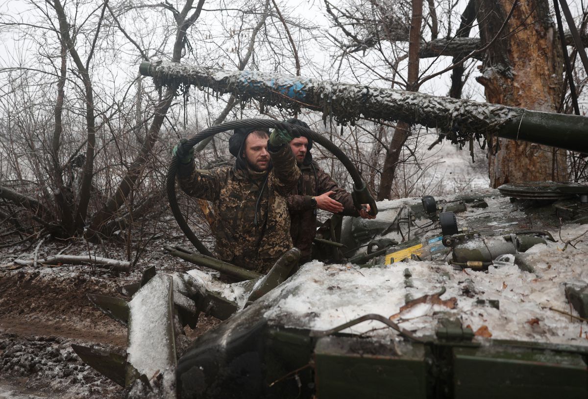 Men in camouflaged uniforms stand beside a tank covered in ice in a snowy, wooded area.