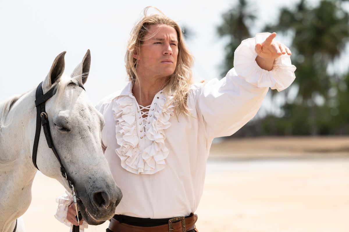 Channing Tatum, in a white ruffled shirt, stands with a white horse on the beach in Lost City and points into the distance.