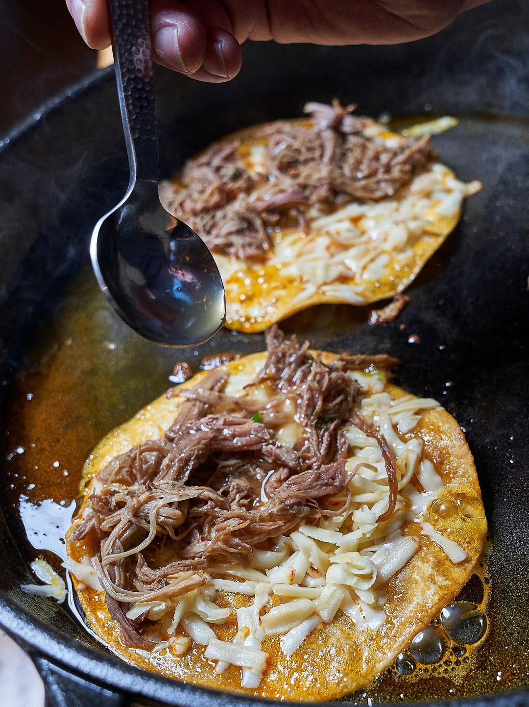 Two birra tacos cook in oil as a spoon drizzles oil into them.