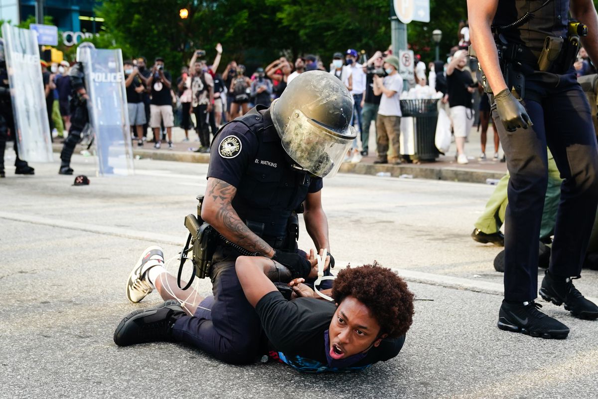 As a black man on the ground cries out, an officer in riot gear handcuffs him.