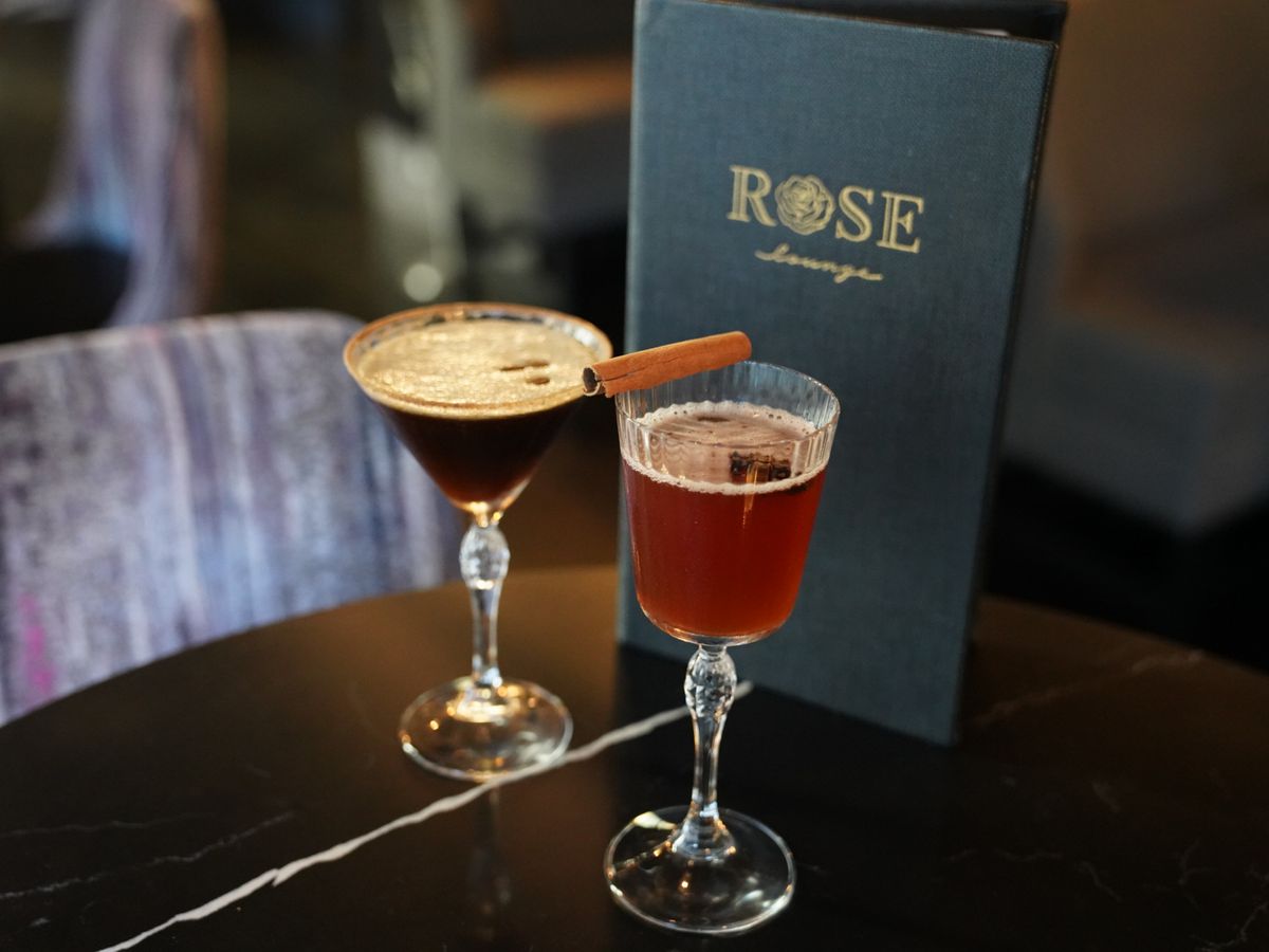 Two cocktails in fancy glasses sit on a black table beside a menu that reads “Rose Lounge.”