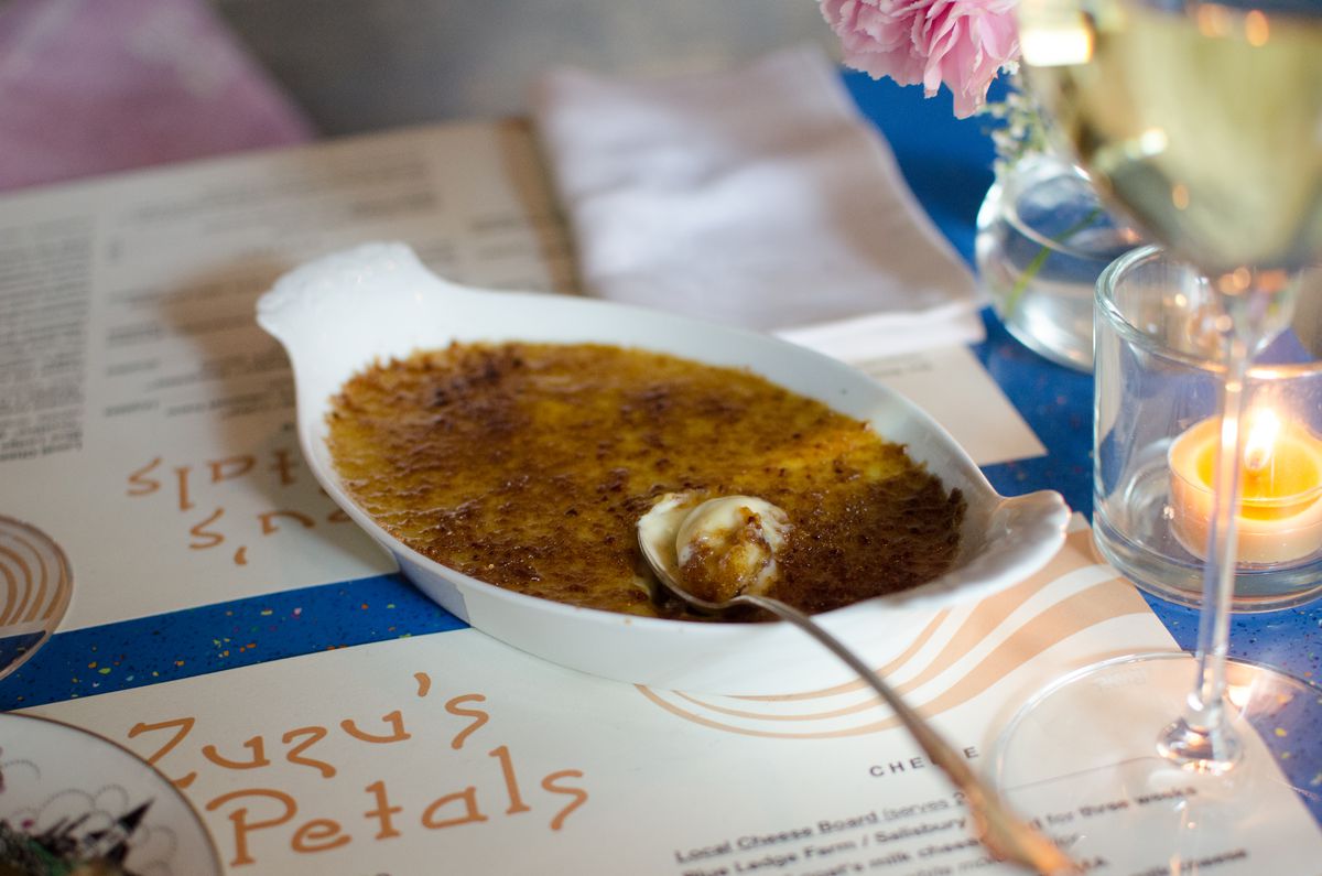 A long spoon breaks into a white oval plate of creme brulee, which sits on a paper menu titled Zuzu’s Petals atop a blue table. There’s a glass of white wine and a small flower vase to the side.