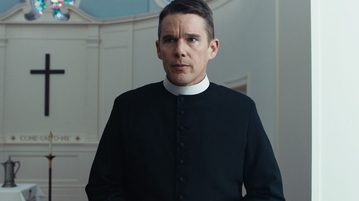 A man in priest attire (Ethan Hawke) stands behind a lectern with a black cross visible in the background.