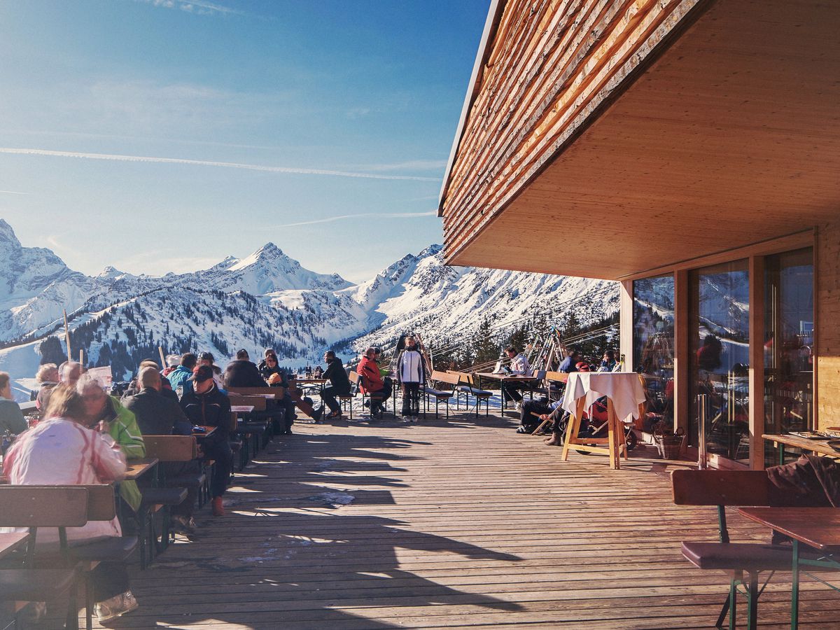 Diners sit at wooden tables on a large deck overlooking sunny snow-covered mountains.