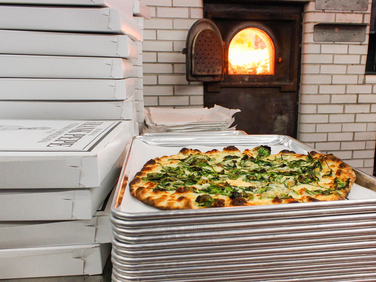 A New Haven-style pizza with a charred crust sits on a stack of metal trays, next to a stack of pizza boxes, in front of a lit pizza oven