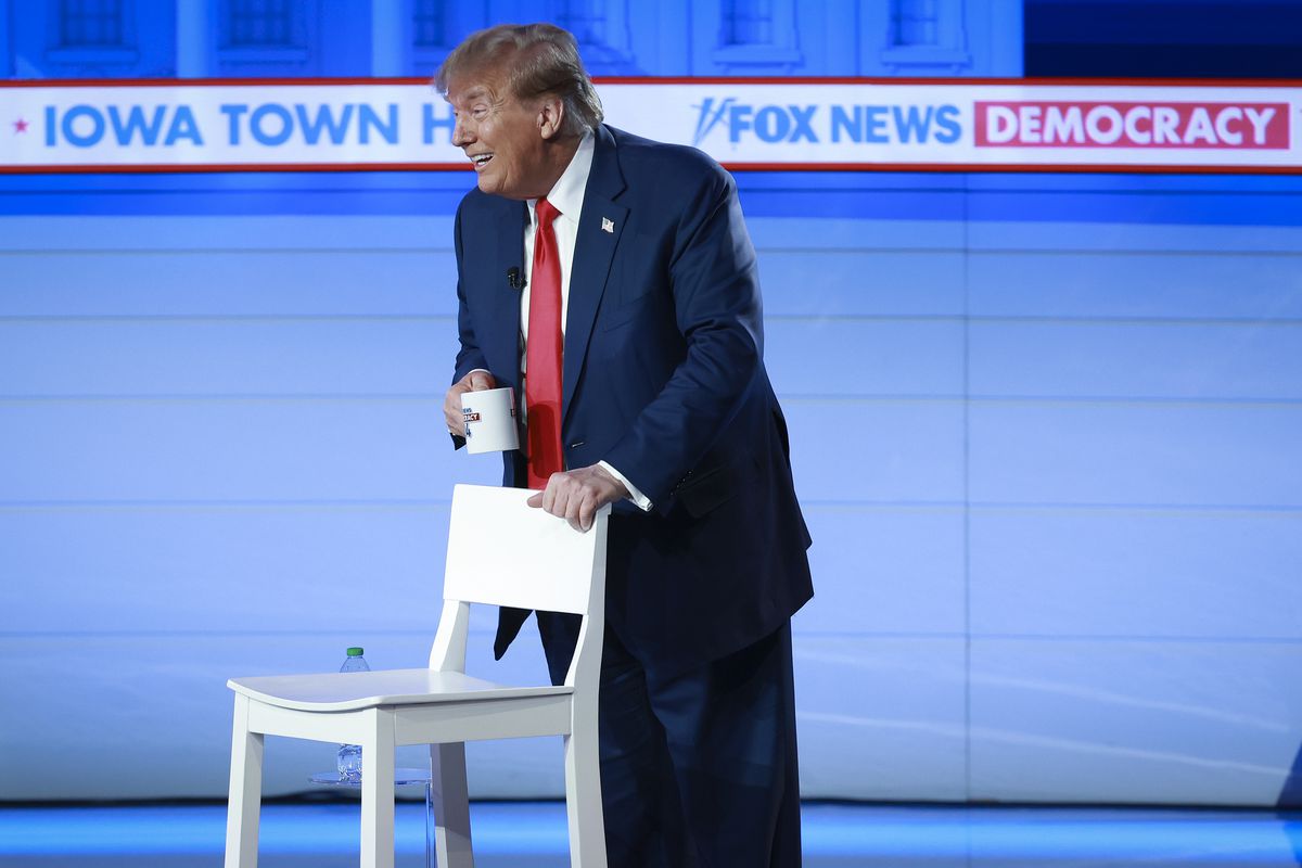 Trump stands onstage leaning on a chair, holding a coffee mug. 