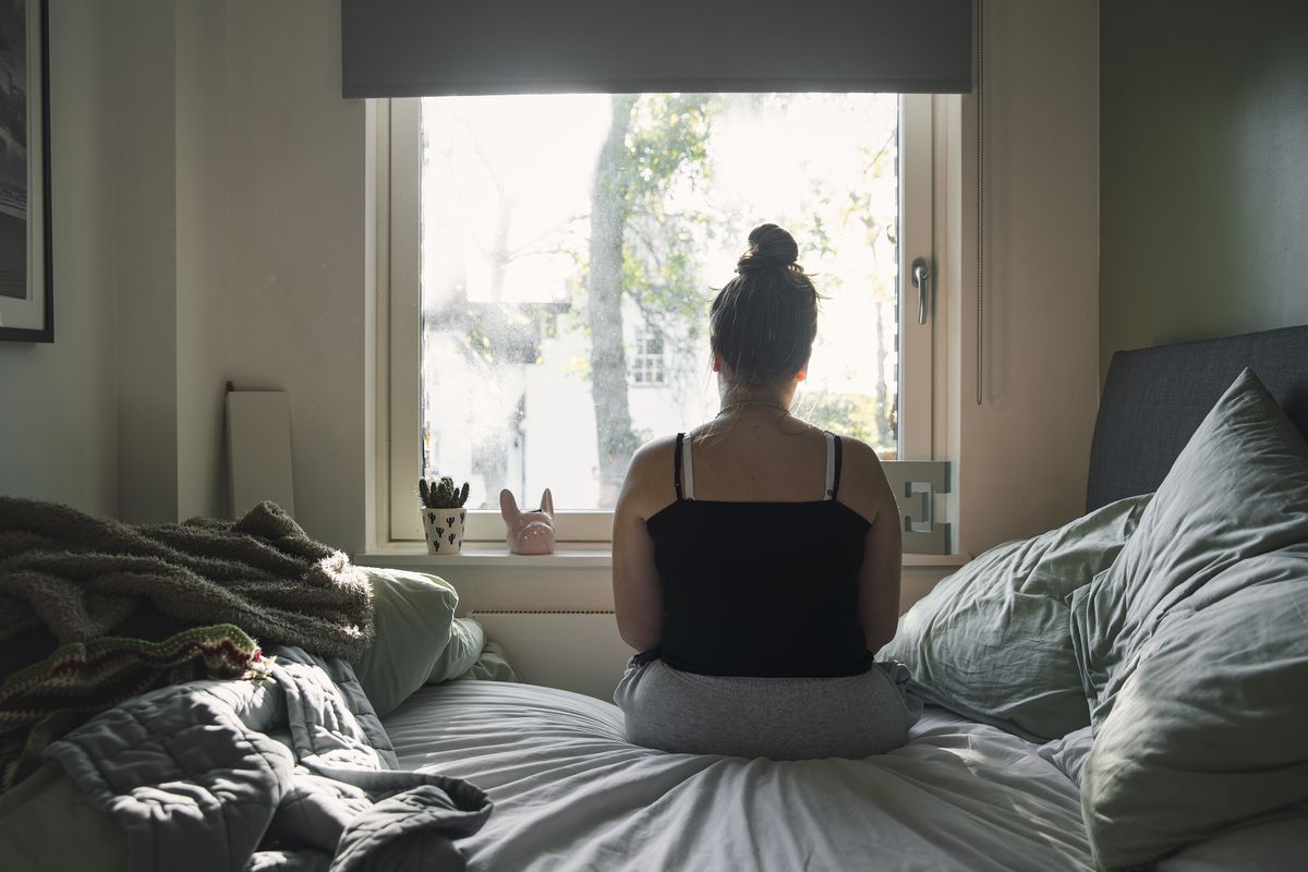 A person sitting on an unmade bed, looking out the window.
