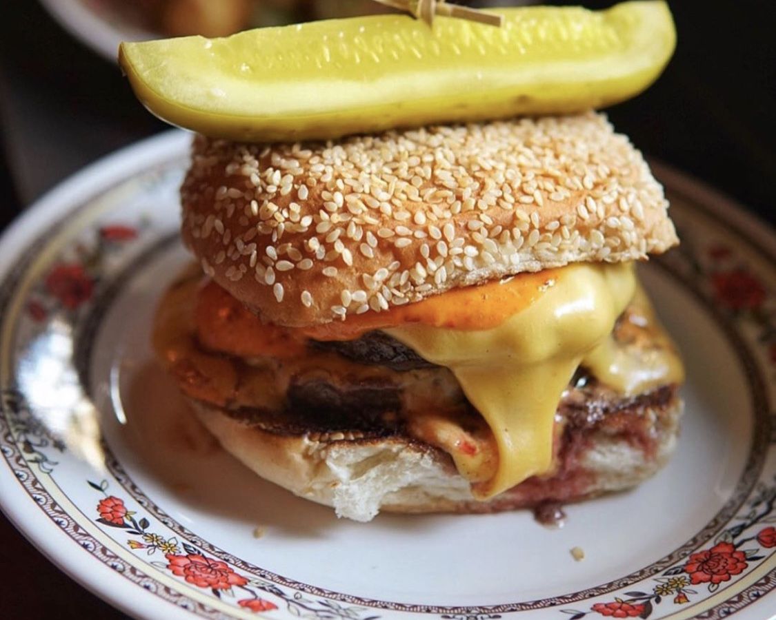 A cheese burger placed on a white plate with a pattern along the border. The cheese burger is sandwiched in a sesame bun with a giant sliced pickle on top held together by a toothpick.