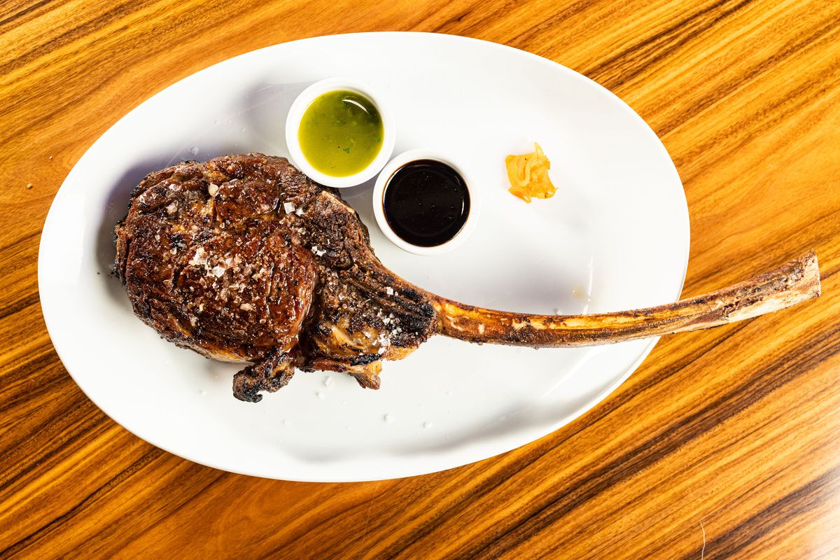 A tomahawk steak sits on a large white plate, bone-in. Next to it are two sauces.