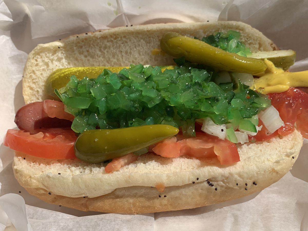 A hot dog in a bun dressed with tomatoes, peppers, onions, and relish on white paper.