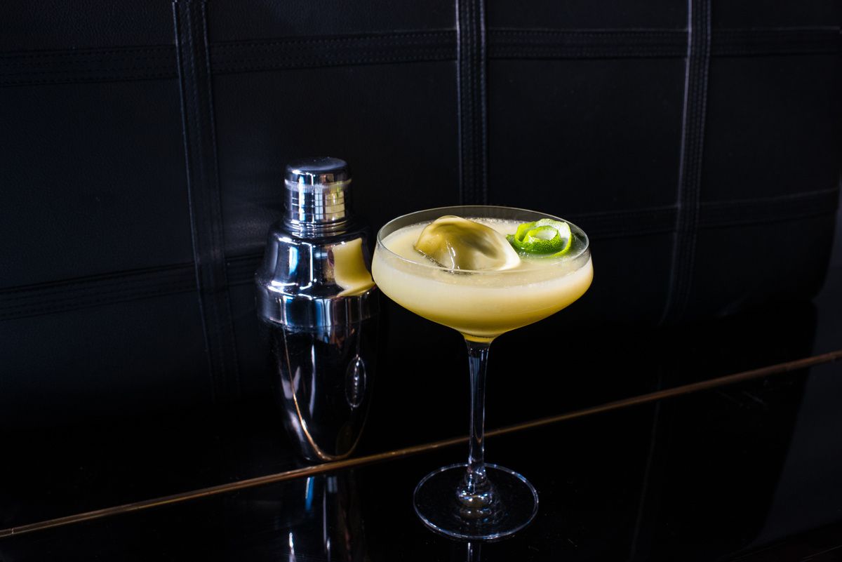A cloudy yellow cocktail in a daiquiri glass against a very black room.