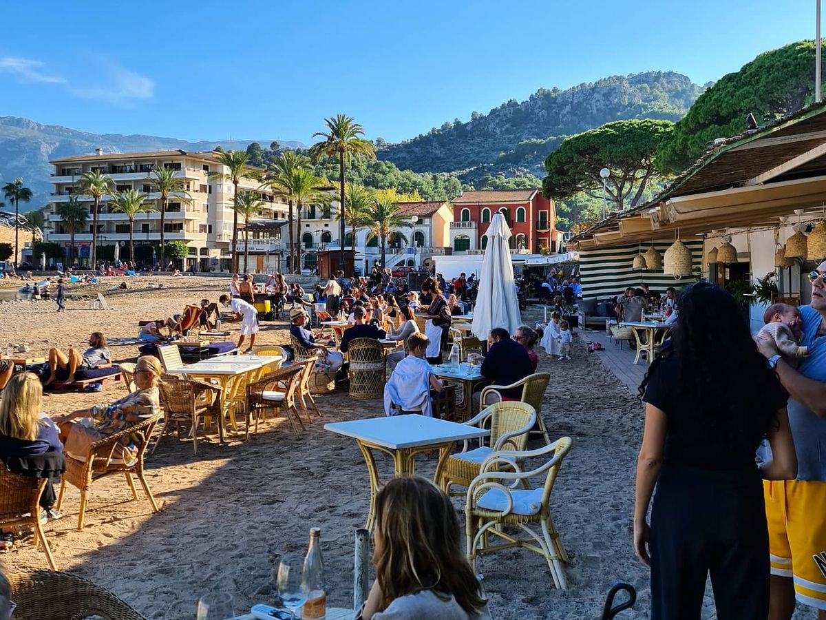 Outdoor diners at tables on the sand, with the hills and buildings of Mallorca stretching out in the distance
