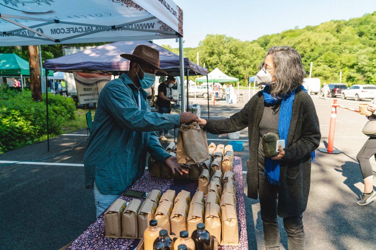 Carvajal hands a bag of coffee to a customer from behind his table at an outdoor farmer’s market.