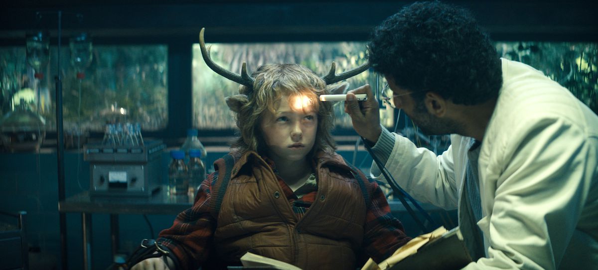 Dr. Singh (Adeel Akhtar) shines a penlight at Gus (Christian Convery), an apprehensive young boy with the antlers and ears of a deer in Sweet Tooth.