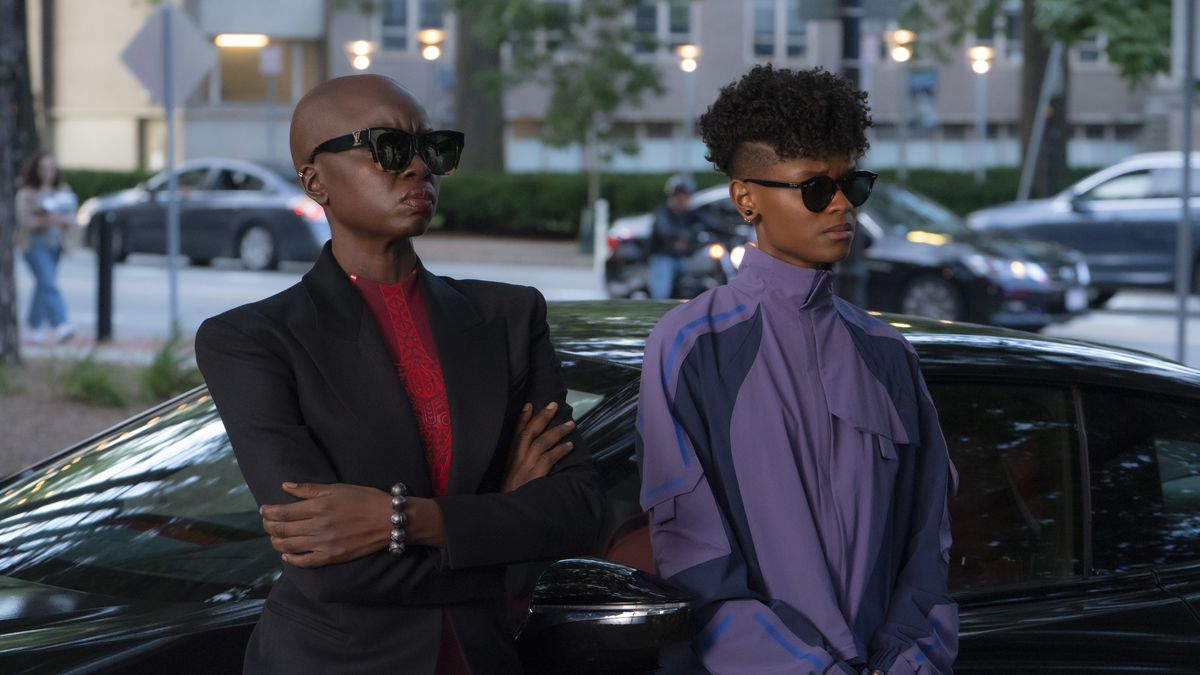 Two women wearing black sunglasses, one in a black suit jacket over a red jumpsuit (Danai Gurira) and the other in a purple techwear jacket (Letitia Wright), lean against a black car with tinted windows.