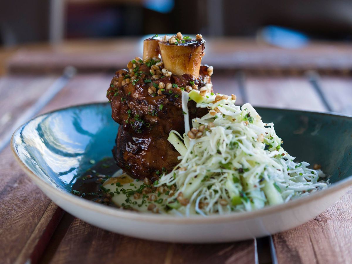 A towering pork shank beside a heap of slaw on a curved ceramic plate on a wooden table