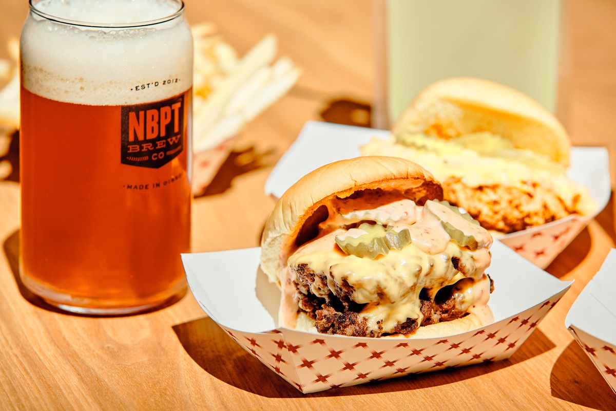 A burger with two patties, cheese, and pickles sits in a paper takeout container next to a glass of beer.