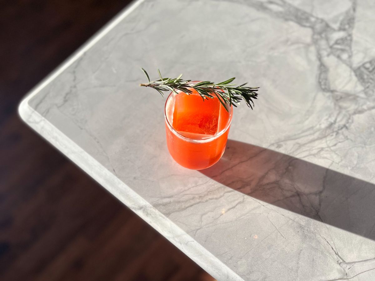 Red orange cocktail with one large block of ice and a sprig of rosemary. 