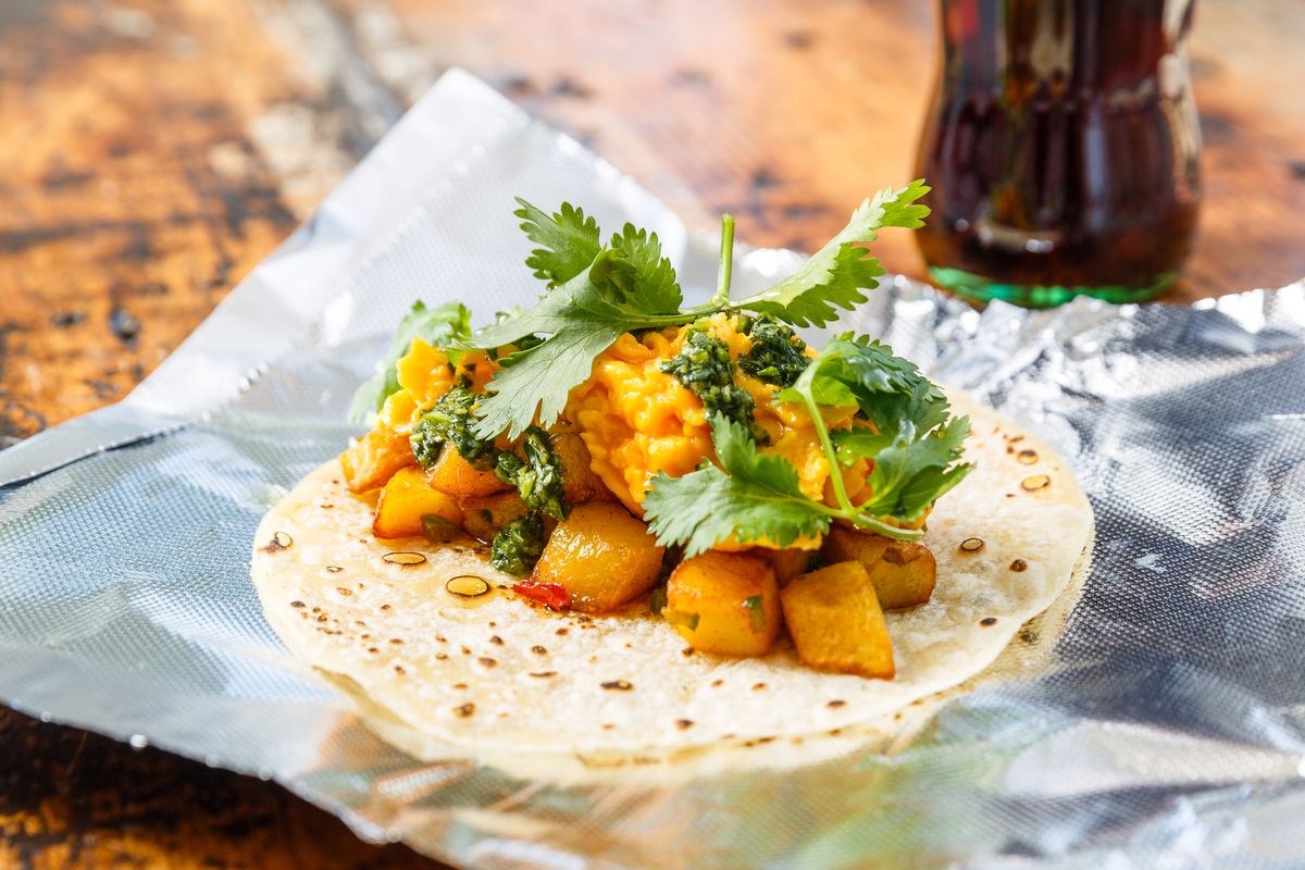 A white flour tortilla filled with cubes of potato, scrambled egg, and green cilantro laid on a foil wrapper.