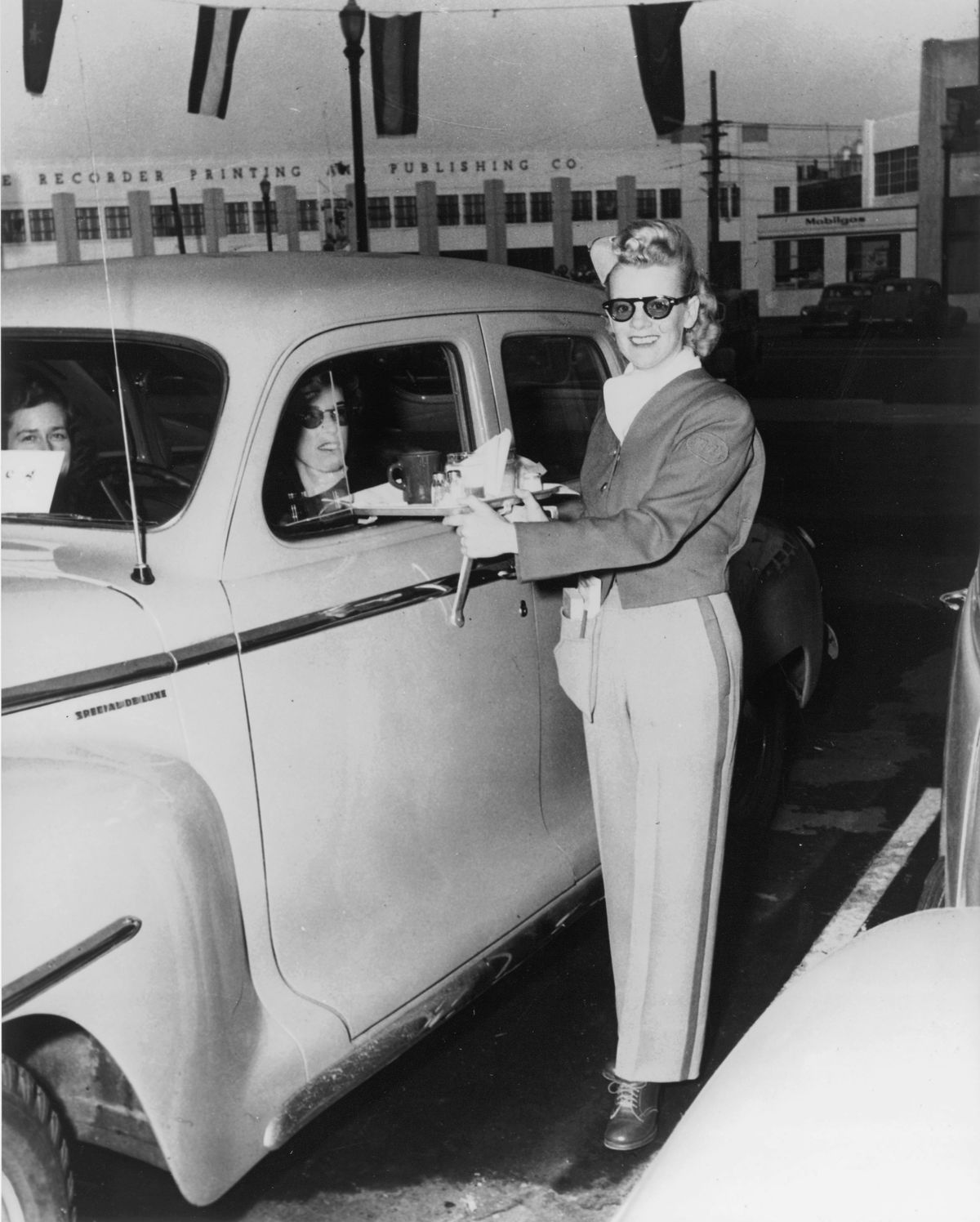 A black and white photo showing an old carhop experience where customers eat alongside their cars.