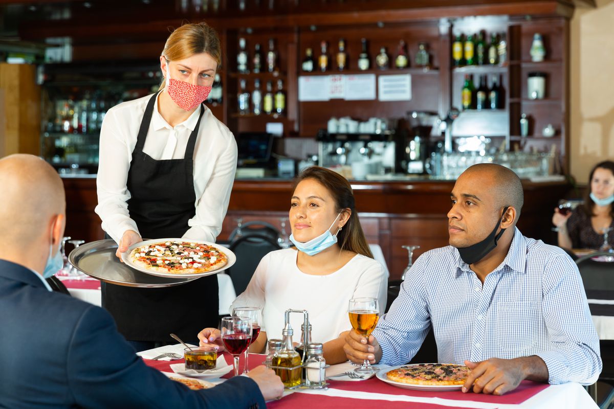 A waitress with a mask dropping off pizza to a table of four.