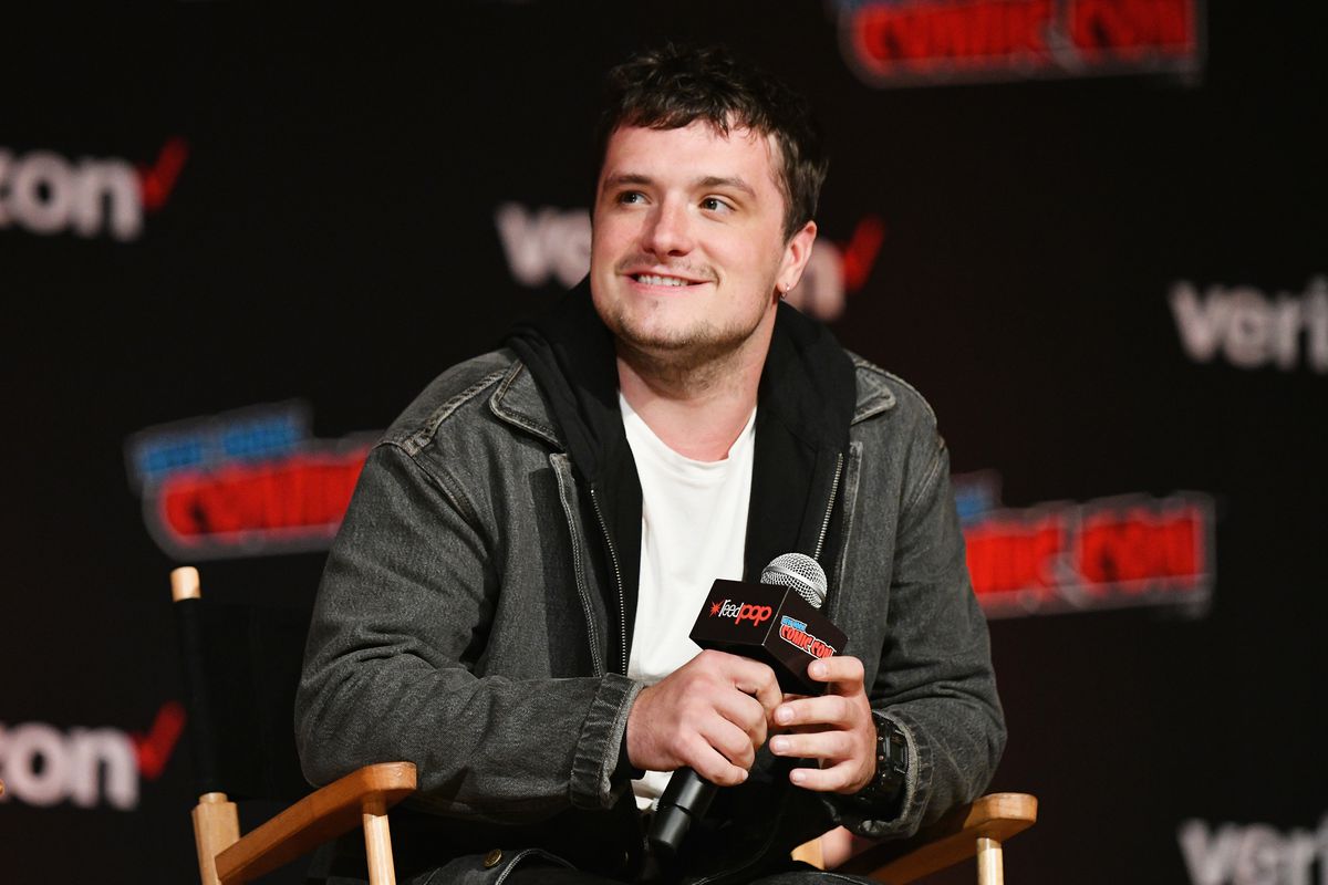 A photo of Josh Hutcherson sitting in a chair while holding a mic on stage. He’s smiling as he looks off to the side. 
