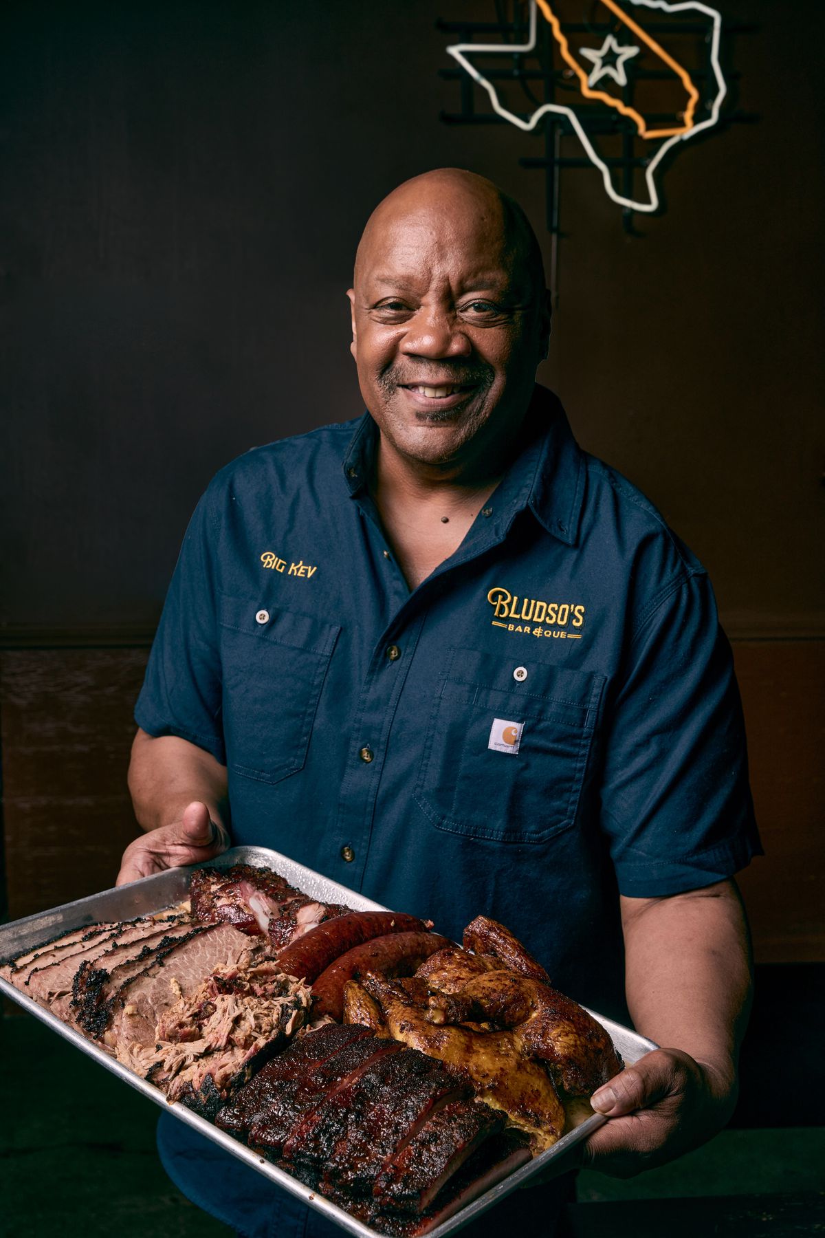 A bald Black man holds a metal tray of barbecue, with a smile.