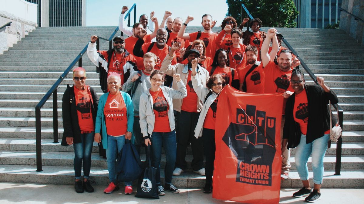 A group portrait of the Crown Heights Tenants Union standing on a concrete staircase. holding a banner There are about two dozen people all wearing red shirts and raising their fists.
