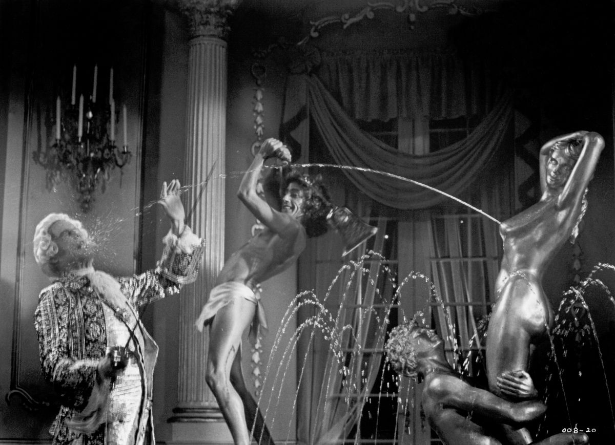 Hoo boy, where to start? In a B&amp;W promo still from Lisztomania, a man in 19th-century court garb and wig stands to the left of the screen, head back, mouth open. A woman covered in shiny paint at the right side of the screen, pretending to be part of a fountain statue, squirts milk from one breast in an arc through the air, into the man’s mouth. The Who’s Roger Daltrey, wearing only a loincloth, stands between them, making a hoop from his arms for the milk to arc through. Wild.