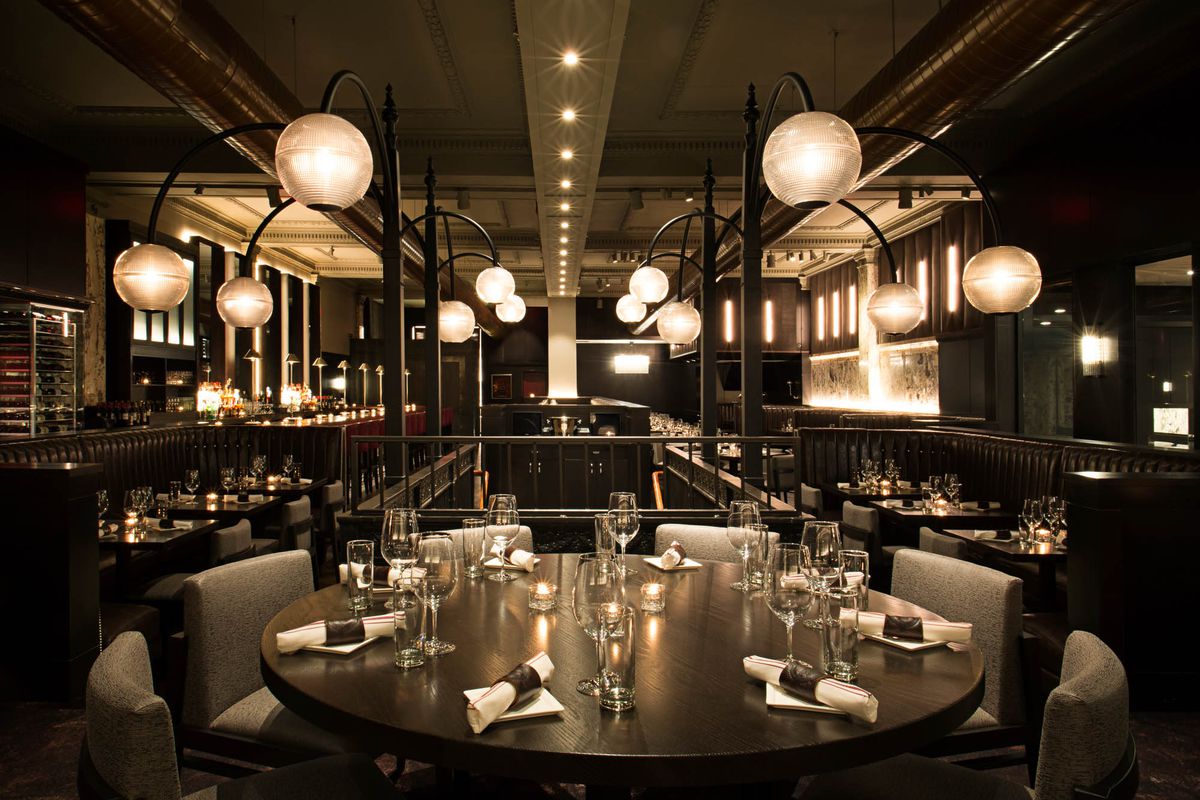 A wide shot of Boston Chops DTX, looking down the length of the restaurant and towards the front door, with elegant light fixtures and a long bar in view