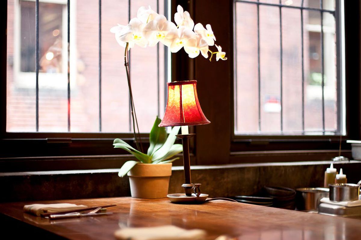 A wooden restaurant table is embellished with a potted white orchid and a small lamp with a red shade.