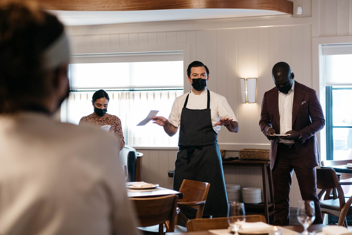 A man in a white shirt, long black apron, and black face mask addresses a room. Next to him stands a man wearing a burgundy suit and white shirt. 