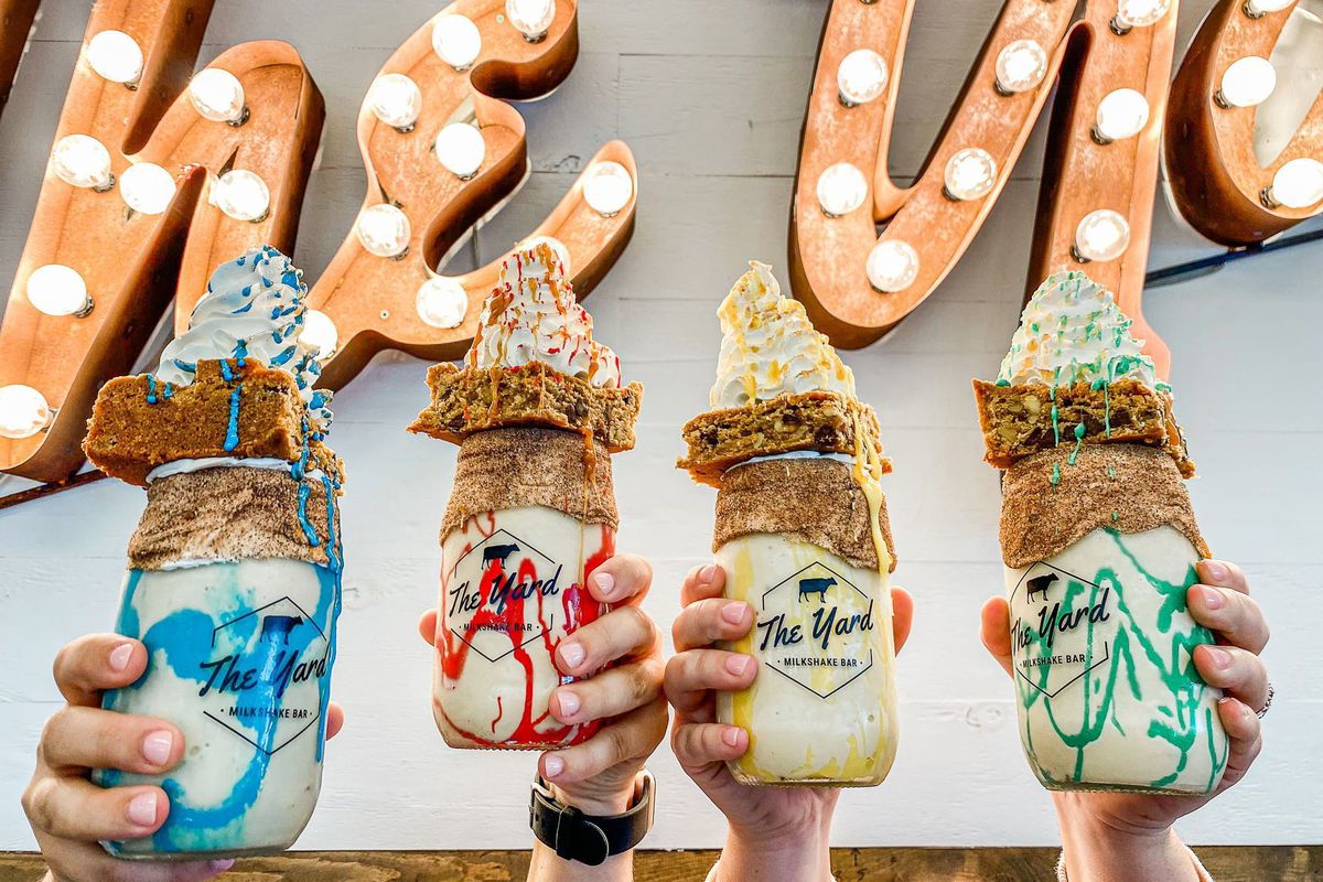 Four hands hold up mason jars filled with milkshakes swirled with colors like yellow, green, red, and blue, topped with blondies and swirls of whipped cream.