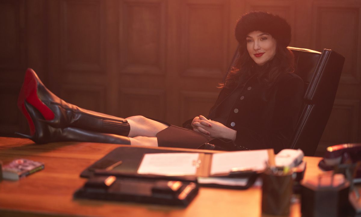 Gal Gadot, in a black fur hat, black jacket, and knee-high black boots, smirks and rests her feet on a desk in Red Notice