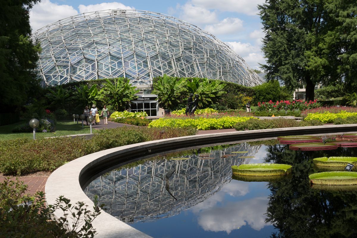 The exterior of the Missouri Botanical Garden. The building is glass with a dome shape. There is a pool in the foreground surrounded by plants. 