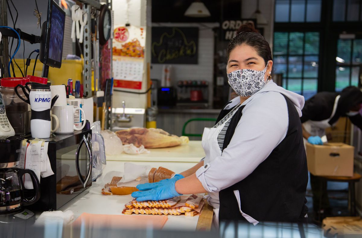 A female staff member of Country Meat Market smiles behind her paisley face mask as she slices bacon to sell at the stall inside the Municipal Market of Atlanta, Sweet Auburn Curb Market. 