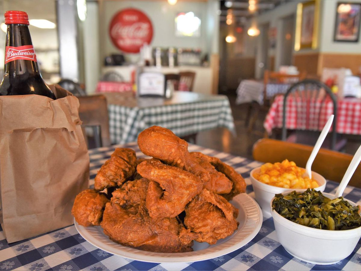 A plate on fried chicken next to two bowls with mac and cheese and greens, next to a bottled beverage in a brown paper bag on a table with blue-and-white-checkered tablecloth.